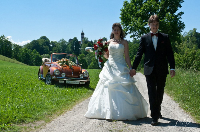 Brautpaar Händchen haltend auf einem Feldweg mit Käfer Cabriolet im Hintergrund